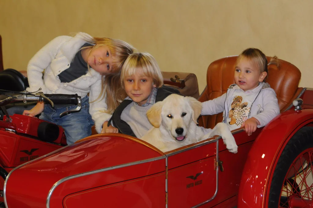 Three beautiful blonde children riding a red Guzzi motorcycle with a Labrador puppy — by Hollyowl Luxury Dog Vests.